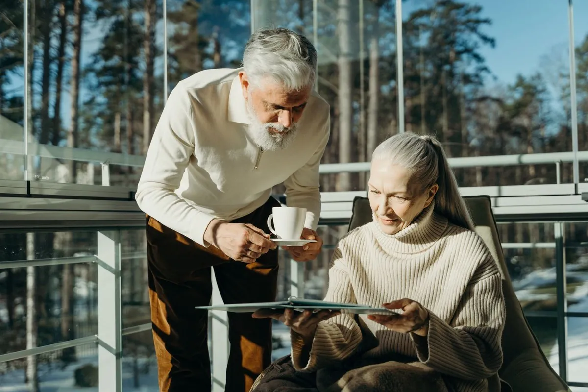 Un homme apporte une tasse de café à sa femme, installée dans un fauteuil près d'une grande fenêtre